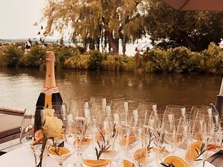 Kranenborgh - Getränke Ein gedeckter Tisch mit Sektflasche, Gläsern mit Orangen und Rosmarin vor einem Fluss im Freien.