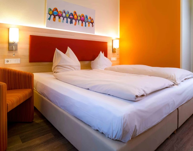 Doppelzimmer im Hotel am Stadtpark Zwei helle Einzelbetten, orangefarbene Akzente im Zimmer, inklusive farbenfrohem Vogelbild an der Wand.