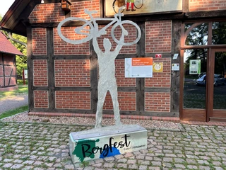 Skulptur Bergfest Eine Skulptur eines Radfahrers, der ein Fahrrad hochhält, vor einem Fachwerkgebäude auf Kopfsteinpflaster.