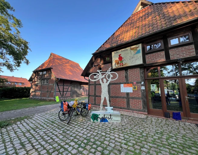 Skulptur Bergfest Skulptur eines Radfahrers mit erhobenem Fahrrad vor einem Fachwerkgebäude mit Fahrradständer.