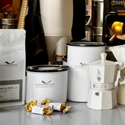Produkte der Elstermühle Kaffeerösterei - Regionale Produkte aus Leipzig Auf einer Auslage vor Kaffeeröstmaschinen stehen die verschiedenen Produkte der Elstermühle Kaffeerösterei aus Leipzig, darunter frische verpackte Kaffeebohnen, Aufbewahrungsbehälter und Espressokocher