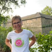 Lachwanderung mit Claudia Thienenkamp, Langenfeld Claudia Thienenkamp, lächelnd im Freien, vor einem steinernen Gebäude in Langenfeld, am Wasser.