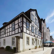 Haus_des-Gastes_2019_Blickrichtung-Marktplatz.png Fachwerkhaus am Marktplatz vor blauem Himmel; nebenan ein Café mit bunten Sonnenschirmen.