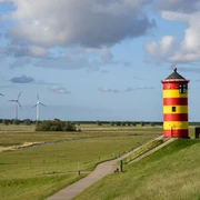 Pilsumer Leuchtturm  Wiesen, Windräder im Hintergrund, grüner Deich mit dem rot-gelben Leuchtturm von Pilsum, blauer Himmel mit Wolken