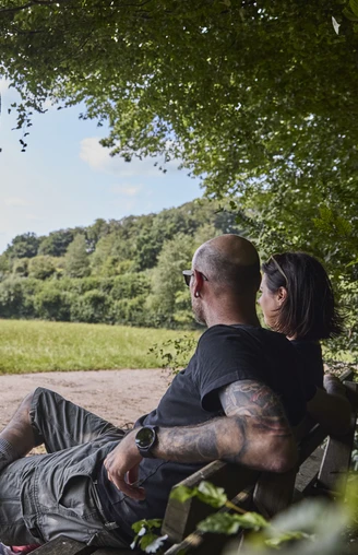 Ein Paar sitzt auf einer Bank am Waldrand und blickt über eine weitläufige Wiesenlandschaft.