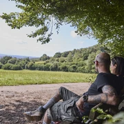 Dörentrup-NABULehrpfad-Teutoburger Wald-LTMGmbH-HirschmeierMediaGmbH&CoKG_025.jpg Ein Paar sitzt auf einer Bank am Waldrand und blickt über eine weitläufige Wiesenlandschaft.