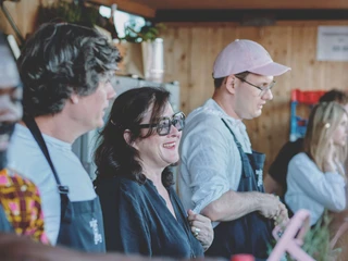 Wine festival on the Rhine In einem gemütlichen Pavillon stehen mehrere Menschen hinter einer Theke und schenken Wein aus. Eine Frau mit Sonnenbrille lächelt fröhlich und unterhält sich mit einer Person, die nicht im Bild zu sehen ist. Blauer Himmel und Holzverkleidung vermitteln eine entspannte Stimmung.Smiling people in aprons serve wine behind a wooden festival booth, sharing friendly interactions.