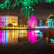 Christmas Garden Markkleeberg Das Bild zeigt eine farbenfrohe, nächtliche Szenerie mit festlich beleuchteten Bäumen und einem historischen Gebäude, das sich in einem ruhigen Gewässer spiegelt. Die intensiven Lichter und Farben verleihen der Umgebung eine magische Atmosphäre.