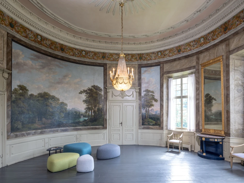 Gartensaal_Copyright_LVR-ZMB, Stefan Arendt 2024.jpg Eleganter, runder Saal mit historischen Wandmalereien, imposantem Kronleuchter und modernen Sitzhockern.