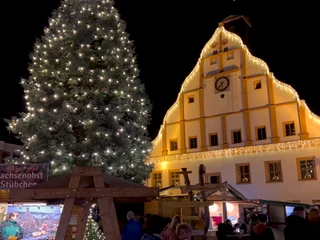 Ein Fest der Sinne - Grimmaer Weihnachtsmarkt  Zu sehen ist das Grimmaer Rathaus weihnachtlich beleuchtet. Im Vordergrund steht ein großer geschmückter Weihnachtsbaum und Stände mit Spezialitäten.