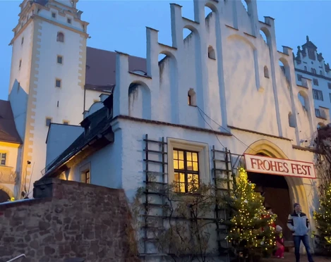 Colditzer Märchenschlossweihnacht Zusehen ist der Eingang von Schloss Colditz. Am Eingangstor sind zwei geschmückte Weihnachtsbäume aufgestellt und über der Tür hängt ein Banner mit der Aufschrift "Frohes Fest"