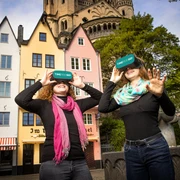 TimeRide VR Old Town Tour Zwei lächelnde Personen mit VR-Brillen vor bunten Stadthäusern und einer Kirche.Two smiling people with VR glasses in front of colorful townhouses and a church.