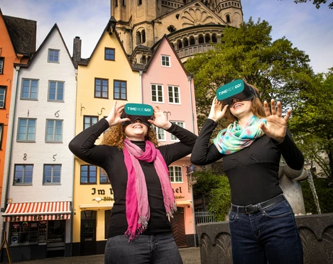 TimeRide VR Old Town Tour Zwei lächelnde Personen mit VR-Brillen vor bunten Stadthäusern und einer Kirche.Two smiling people with VR glasses in front of colorful townhouses and a church.