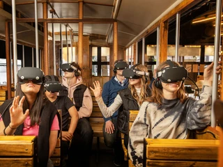 TimeRide SENSEUM - Virtual time travel through Cologne Sechs Personen in einem historischen Straßenbahnwagen, die Virtual-Reality-Headsets tragen und dabei in verschiedene Richtungen blicken. Die Passagiere wirken fasziniert und neugierig, während sie in eine virtuelle Welt eintauchen:Six people in a historic streetcar carriage wearing virtual reality headsets and looking in different directions. The passengers appear fascinated and curious as they immerse themselves in a virtual world: