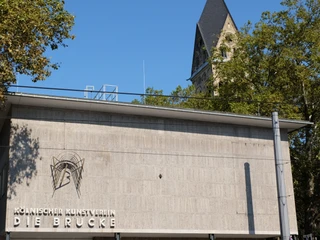 Kölnischer Kunstverein Der Kölnische Kunstverein: Moderne Betonarchitektur vor einem Kirchturmhintergrund, von Bäumen umrahmt.The Kölnischer Kunstverein: Modern concrete architecture in front of a church tower background, framed by trees.
