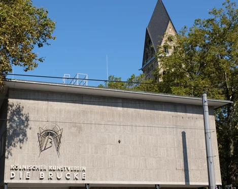 Kölnischer Kunstverein Der Kölnische Kunstverein: Moderne Betonarchitektur vor einem Kirchturmhintergrund, von Bäumen umrahmt.The Kölnischer Kunstverein: Modern concrete architecture in front of a church tower background, framed by trees.
