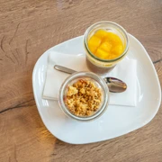 Café Bös Frühstücksteller, Ratingen Zwei Gläser auf einem weißen Teller, gefüllt mit frischem Obst und knusprigem Crumble, präsentiert.