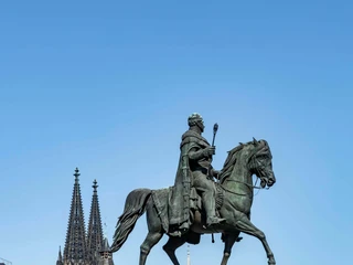Equestrian statue of King Frederick William III. Das Bild zeigt das bronzene Reiterstandbild von König Friedrich Wilhelm III. auf einem steinernen Sockel. Der detailreich gearbeitete König sitzt mit stolzer Haltung auf einem majestätischen Pferd. Entlang des Sockels sind ebenfalls bronzene Figuren abgebildet, die bedeutende Persönlichkeiten jener Epoche darstellen. Im Hintergrund sind die beiden markanten Türme des Kölner Doms erkennbar. The picture shows the bronze equestrian statue of King Frederick William III on a stone pedestal. The richly detailed king sits proudly on a majestic horse. Along the pedestal are also bronze figures depicting important personalities of the era. The two striking towers of Cologne Cathedral can be seen in the background.