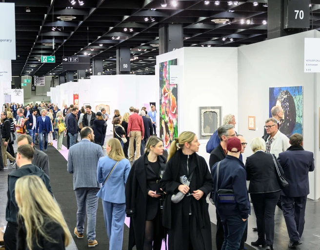 ART COLOGNE Menschen flanieren entlang der Stände, betrachten aufmerksam verschiedene Kunstwerke, die an weißen Wänden präsentiert werden. Im Vordergrund zieht eine bunte Mischung aus Gemälden die Blicke der Betrachter*innen auf sich, während die Decke durch industrielle Beleuchtung hervorsticht. Die Szene wirkt lebendig und zeigt das Interesse der Menschen an zeitgenössischer Kunst.People stroll along the stands, attentively looking at various works of art presented on white walls. In the foreground, a colorful mixture of paintings catches the viewer's eye, while the ceiling stands out with industrial lighting. The scene looks lively and shows people's interest in contemporary art.