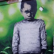 Mural Ein großformatiges Graffiti zeigt einen jungen Jungen mit ernstem Blick vor einem grünen Hintergrund. Auf seinem Pullover steht die verblasste Botschaft: „I WILL NEVER SAY WHAT YOU WANT TO HEAR“.A large-format graffiti shows a young boy with a serious look in front of a green background. The faded message on his sweater reads: "I WILL NEVER SAY WHAT YOU WANT TO HEAR".