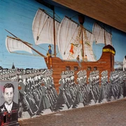 Mural Ein großflächiges Wandgemälde zeigt eine historische Szene der Edelweißpiraten in Köln. Ein Segelschiff mit weißen Segeln zieht über den Ozean, während im Vordergrund eine marschierende Menschenmenge zu sehen ist. Am linken Bildrand ist ein Porträt eines jungen Mannes zu erkennen. Das lebendige, historische Kunstwerk ziert eine Unterführung und erinnert an die soziale Geschichte der Stadt.A large-scale mural shows a historical scene of the Edelweiss Pirates in Cologne. A sailing ship with white sails is sailing across the ocean, while a marching crowd can be seen in the foreground. A portrait of a young man can be seen on the left-hand edge of the picture. The lively, historical work of art adorns an underpass and is a reminder of the city's social history.