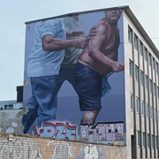 Mural Das Bild zeigt ein großflächiges Wandgemälde auf einem mehrstöckigen Gebäude. Zwei männliche Figuren sind in einer dynamischen Bewegung dargestellt, wobei eine Person ein blaues T-Shirt trägt und die andere, halbnackte, von dieser gestützt wird.The picture shows a large-scale mural on a multi-storey building. Two male figures are depicted in a dynamic movement, with one person wearing a blue T-shirt and the other, half-naked, being supported by it.