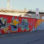 Mural Eine lange, leuchtend rote Mauer am Straßenzug ist mit bunten, abstrakten Kunstwerken verziert.A long, bright red wall along the street is decorated with colorful, abstract works of art.