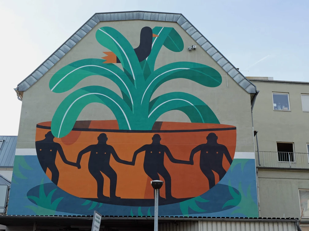 Mural Das Wandgemälde auf einer Hauswand zeigt vier figürlich stilisierte Personen in einer großen, pflanzenähnlichen Schale, umgeben von grünen und orangefarbenen Formen.The mural on a house wall shows four stylized figures in a large, plant-like bowl, surrounded by green and orange shapes.