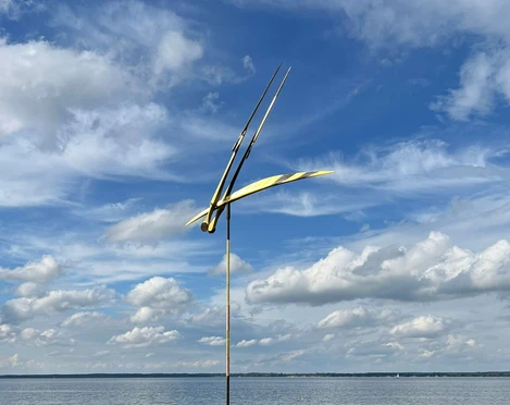 Skulptur vor der Promenade Libelle im Wind Skulptur mit filigranen Flügeln schwebt über ruhigem Wasser vor wolkigem Himmel.