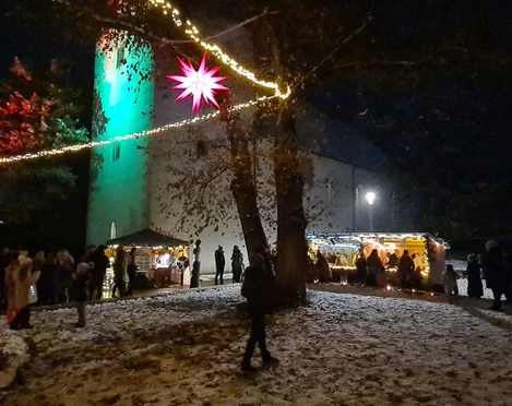 Löbnitzer Adventsmarkt Das Bild zeigt einen stimmungsvoll beleuchteten Weihnachtsmarkt bei Nacht, mit schneebedecktem Boden, geschmückten Verkaufsständen und einem markanten roten Stern, der in der Mitte zwischen den Bäumen hängt.