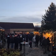 Löbnitzer Weihnachtsmarkt Das Bild zeigt einen Weihnachtsmarkt in der Dämmerung, mit Menschen, die sich um festlich geschmückte Stände und einen großen, beleuchteten Weihnachtsbaum versammeln, während die Lichter und Dekorationen eine gemütliche Atmosphäre schaffen.