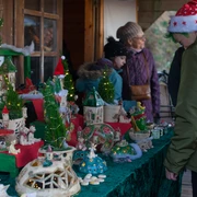 Schkeuditzer Weihnachtsmarkt Das Bild zeigt einen Stand auf einem Weihnachtsmarkt, der handgemachte Dekorationen und Miniaturhäuser ausstellt, festlich dekoriert mit kleinen Weihnachtsbäumen und Lichterketten, während Besucher in Winterkleidung die ausgestellten Objekte interessiert betrachten.