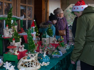 Schkeuditzer Weihnachtsmarkt Das Bild zeigt einen Stand auf einem Weihnachtsmarkt, der handgemachte Dekorationen und Miniaturhäuser ausstellt, festlich dekoriert mit kleinen Weihnachtsbäumen und Lichterketten, während Besucher in Winterkleidung die ausgestellten Objekte interessiert betrachten.