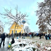 Weihnachtsmarkt Nischwitz  Das Bild zeigt eine winterliche Szene auf einem Weihnachtsmarkt, bei dem zahlreiche Besucher vor einem gelben Gebäude stehen, während Kinder im Schnee spielen.