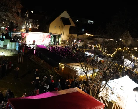 Adventsmarkt Stadt Rötha Das Bild zeigt einen stimmungsvollen Weihnachtsmarkt bei Nacht, mit beleuchteten Marktständen und einem festlich geschmückten Weihnachtsbaum, umgeben von Besuchern, die das Ambiente genießen.