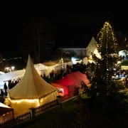 Adventsmarkt Stadt Rötha Das Bild zeigt einen stimmungsvollen Weihnachtsmarkt bei Nacht, mit beleuchteten Marktständen und einem festlich geschmückten Weihnachtsbaum, umgeben von Besuchern, die das Ambiente genießen.