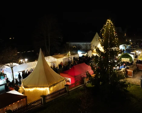 Adventsmarkt Stadt Rötha Das Bild zeigt einen stimmungsvollen Weihnachtsmarkt bei Nacht, mit beleuchteten Marktständen und einem festlich geschmückten Weihnachtsbaum, umgeben von Besuchern, die das Ambiente genießen.