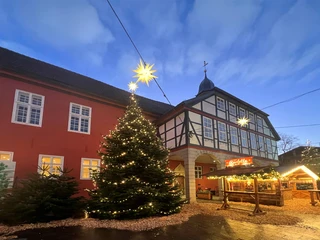 Adventszauber Nienburg (3).jpg Historisches Fachwerkgebäude mit festlich beleuchtetem Weihnachtsbaum und weihnachtlichem Marktstand.