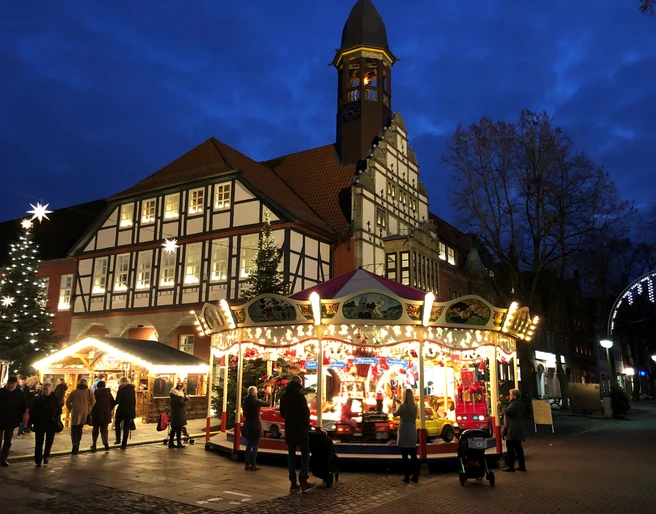 Adventszauber Nienburg (2).jpg Weihnachtlich beleuchteter Marktplatz in Nienburg mit Karussell und Fachwerkgebäude bei Dämmerung.