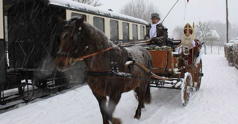 Eine historische Dampflokomotive zieht einen offenen Waggon, geschmückt mit einem Weihnachtsmann und einem Mann in traditioneller Kleidung, durch eine verschneite Winterlandschaft.
