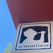 La Vincaillerie Ein quadratisches Schild zeigt das stilisierte Profil einer Person, die aus einem Weinglas trinkt, mit der Aufschrift "la vincaillerie" darunter, auf rotem Gebäudehintergrund vor blauem Himmel.A square sign shows the stylized profile of a person drinking from a wine glass, with the inscription "la vincaillerie" underneath, on a red building background against a blue sky.