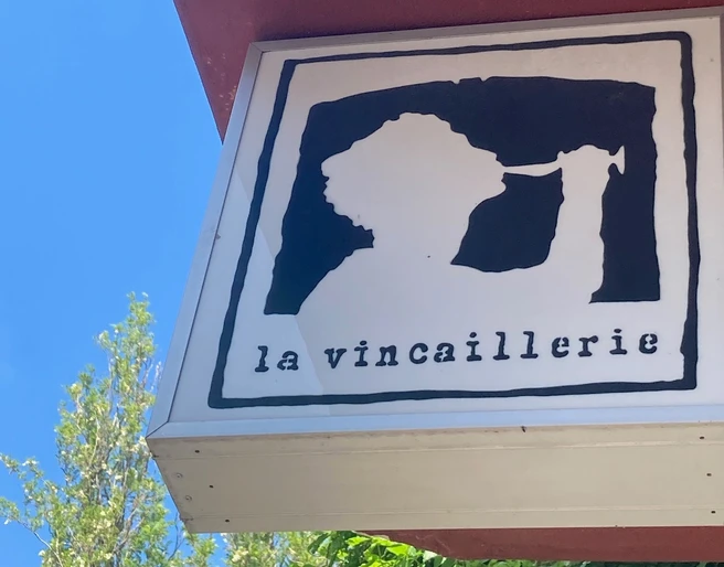 La Vincaillerie Ein quadratisches Schild zeigt das stilisierte Profil einer Person, die aus einem Weinglas trinkt, mit der Aufschrift "la vincaillerie" darunter, auf rotem Gebäudehintergrund vor blauem Himmel.A square sign shows the stylized profile of a person drinking from a wine glass, with the inscription "la vincaillerie" underneath, on a red building background against a blue sky.