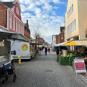 Marktszene in einer gepflasterten Straße mit Ständen voller frischer Produkte und bunten Marktschirmen.