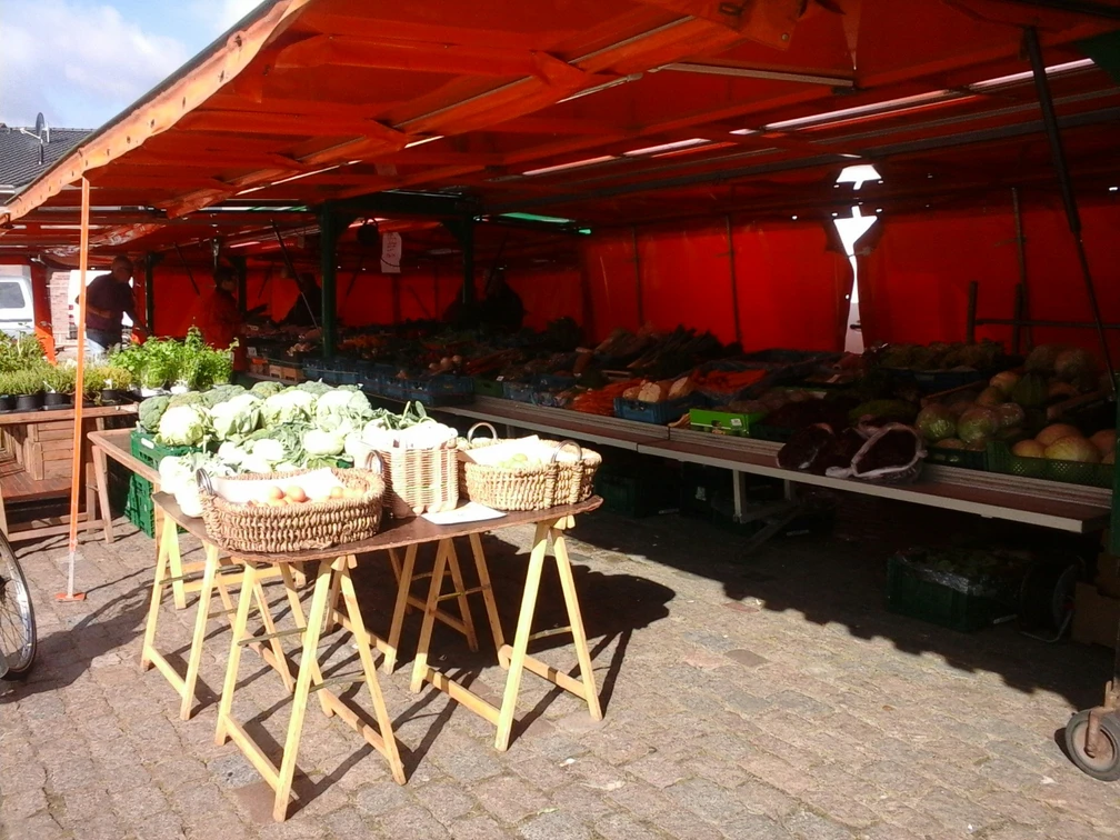 Stand auf dem Aper Wochenmarkt Ein Marktstand mit frischen Gemüsekisten unter orangefarbenen Planen auf einem gepflasterten Platz.