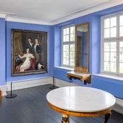 Die Brügelmanns Cromford Ein lichtdurchfluteter Raum mit blauen Wänden, Holzuhr, Gemälde, Spiegel und einem runden Tisch.