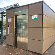 Bahnhof Ratzeburg_WC1_Petra Roemer_20241022.jpg Am Bahnhof steht ein öffentliches Toilettenhäuschen mit silberner Tür, daneben überdachte Fahrradstellplätze