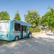 Eiswagen Fred Ein türkisfarbener Eiswagen steht auf einem Gehweg, umgeben von grünen Bäumen und Sträuchern.