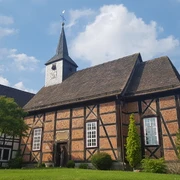 Ein Fachwerkgebäude mit Kirchturm und Wetterfahne vor blauem Himmel und flankiert von Bäumen.