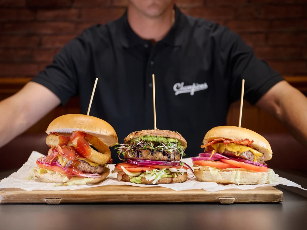 Champions Sports Bar - Gastronomie in Leipzig Ein Koch hält ein Holzbrett, auf dem drei Burger liegen