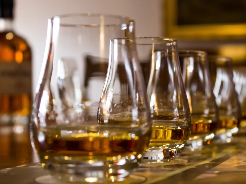 Eine Reihe von Whiskeygläsern auf einem Tisch, im Hintergrund unscharfe Whiskeyflaschen.